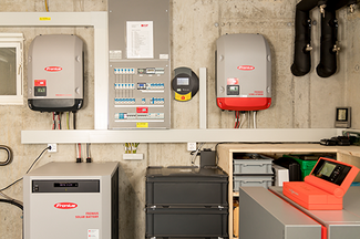 Technikraum mit mehreren an der Betonwand montierten Geräten, darunter zwei Fronius‑Wechselrichter, ein Sicherungs‑ und Verteilerkasten, ein Batteriespeicher sowie sichtbare Kabel, Leitungen und Rohrinstallationen; die Anlage ist übersichtlich und professionell installiert.